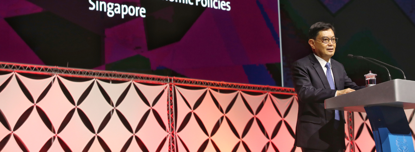 Man in suit with glasses at podium; screen reads 'Singapore Economic Policies'.